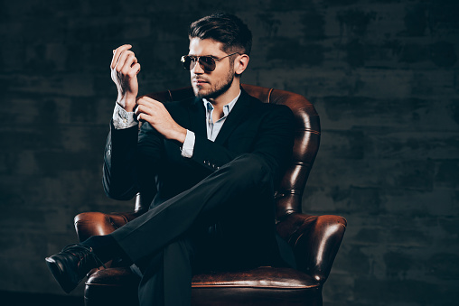 Young handsome man in suit and sunglasses adjusting sleeve on his shirt while sitting in leather chair against dark grey background
