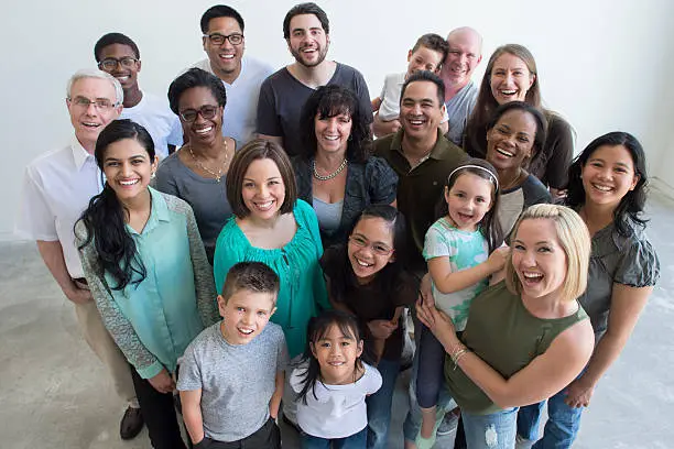 A multi-ethnic group of multi-generational families are standing together happily in a group. They are smiling and looking up at the camera.