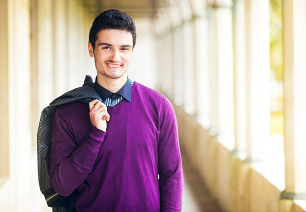An image of a young turkish man standing proud and successful, smiling to the camera. He is wearing trendy youthful purple sweater and is holding an outer jacket thrown over the back. It is a sunny warm picture. An outdoors corridor with columns visible in the background. Copy space available.