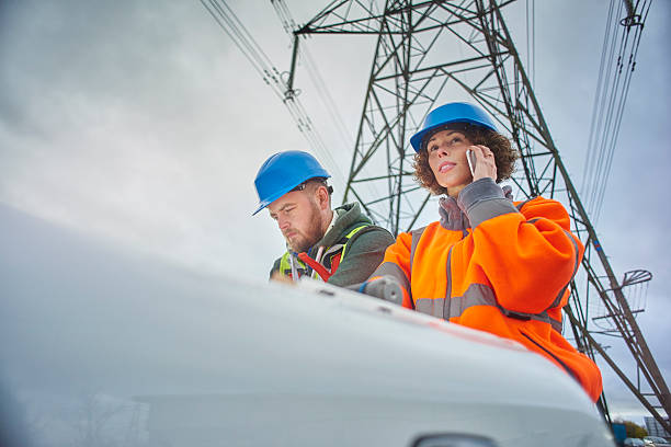 electricity engineers beneath a pylon male and female engineers beneath an electricity pylon looking at plans on the bonnet of their van . The female engineer is on the phone. power cable photos stock pictures, royalty-free photos & images