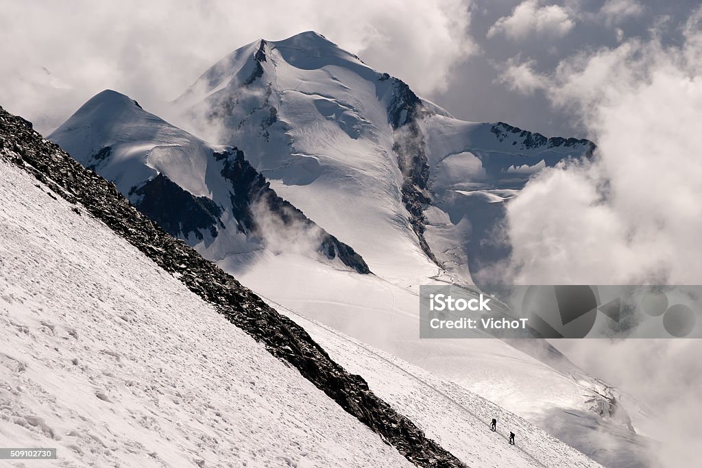 Mountaineers in the distance climbing a steep slope Mountaineering team in the distance climbing the steep slope of Mt Breithorn in Italian Alps. On the background Mt Monte Rosa is rising. Activity Stock Photo