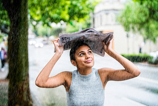 Young Indian woman standing outdoors in the rain while holding newspapers over her head. Sydney, Australia