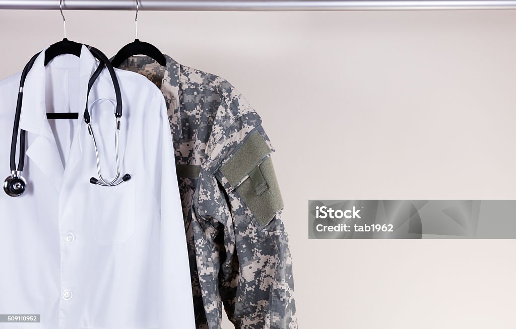 Medical white consultation coat and military uniform on hanger Medical Doctor Consultation white coat, stethoscope around collar, and military uniform hanging on rack. Off white wall in background. Horizontal layout with copy space. Military Stock Photo
