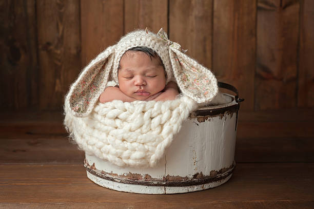 Newborn Girl Wearing a Bunny Bonnet A three week old newborn baby girl sleeping in a little, white wooden bucket. She is wearing a cream colored bunny rabbit bonnet. Shot in the studio on a wood background. babies only photos stock pictures, royalty-free photos & images