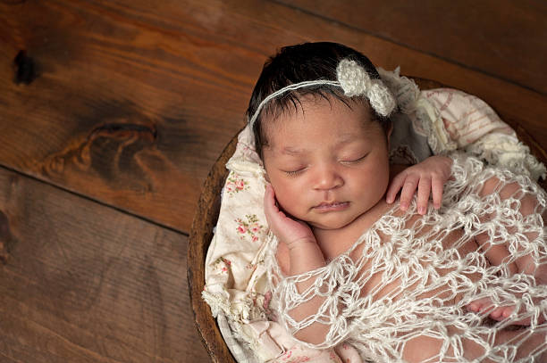 Newborn Girl Sleeping in Wooden Bowl A three week old newborn baby girl sleeping in a little, wooden bowl. She is wearing a cream colored bow headband and swaddled with a decorative wrap. Shot in the studio on a wood background. baby girls stock pictures, royalty-free photos & images