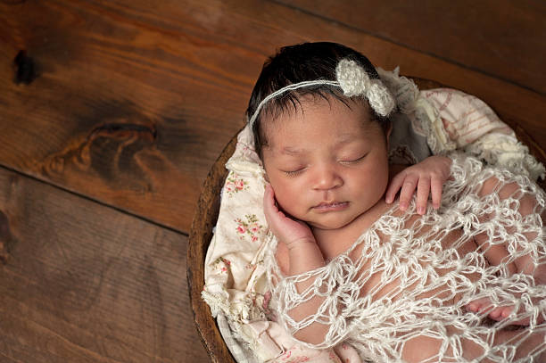 Newborn Girl Sleeping in Wooden Bowl A three week old newborn baby girl sleeping in a little, wooden bowl. She is wearing a cream colored bow headband and swaddled with a decorative wrap. Shot in the studio on a wood background. black shawl wrap stock pictures, royalty-free photos & images