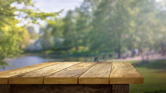 An empty wooden table in a park in the summer. With a shallow depth of focus, a pond, trees, patio and people are all out of focus in the soft background.