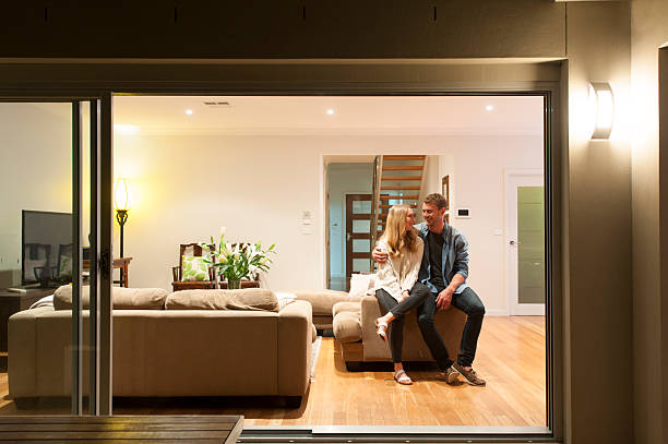 Couple relaxing in their home at night. Couple relaxing in their home at night. They are both wearing casual clothes and embracing. They are looking at each other and smiling. The house is contemporary with an open plan al fresco style. Copy space show home stock pictures, royalty-free photos & images