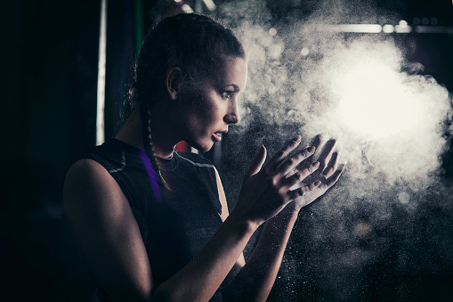 Silhouette of a young fit woman standing in a cross training rack, applying magnesium gym chalk on her hands.