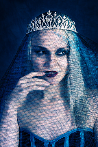 Vertical studio shot on blue of young woman in blue and black corset and blue tulle veil over crystal crown. Front view touching lips looking to viewer's right. Concept of evil queen plotting.