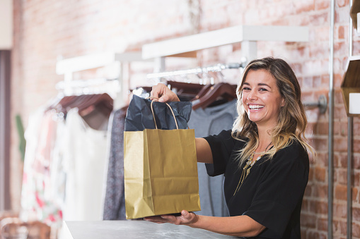 A young woman holding a shopping bag at the checkout counter of a clothing store.  She is smiling at the camera, relaxed and confident.  She may be the owner of this small business, or a cashier or sales assistant.  Racks of clothing on hangers are lined up against the brick wall behind her.