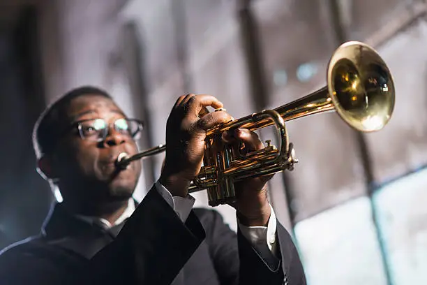 An African American man playing a trumpet.  The musician is wearing a black jacket and tie, performing on stage. It is a low angle view, with the focus on his hands and the musical instrument.