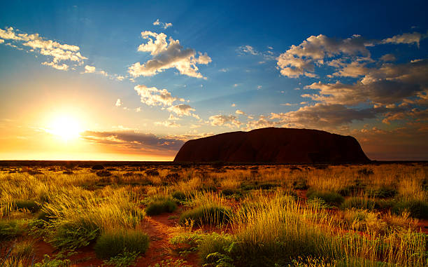 Uluru At Dawn The Northern Territory, Australia - May 18, 2011: The sun rises over the vast open plains of the Australian Outback, illuminating the grass & scrub in the foreground and revealing the silhouetted mass of  Uluru on the horizon. northern territory australia stock pictures, royalty-free photos & images