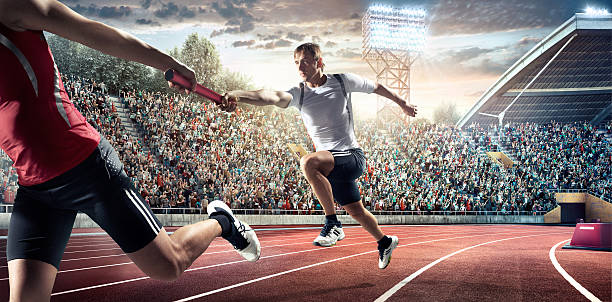 Running Pass on . Stadium A male athletes sprinting and passing race baton on the track on the . stadium. The bleachers are full of spectators. The sky is blue and cloudy. Sportsmen are wearing an unbranded sport uniform. relay photos stock pictures, royalty-free photos & images