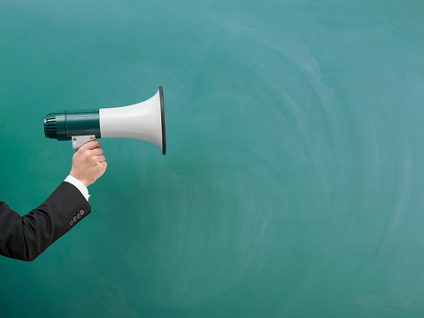 Modern Megaphone In Human Hand On Green Blank Blackboard.The megaphone is on the left side of image.It is in human hand.Studio shot.Shot with medium format camera Hasselblad in studio.Intentional slight shadow of megaphone for real look of image.Model is wearing a black suit.