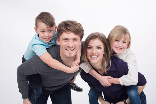 A mother and father are giving their children a piggy back ride. They are all smiling while looking at the camera. The picture is isolated on white.