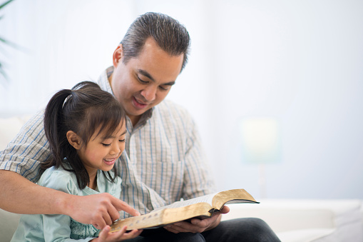 A father is reading the Bible to his daughter on the couch in their home.