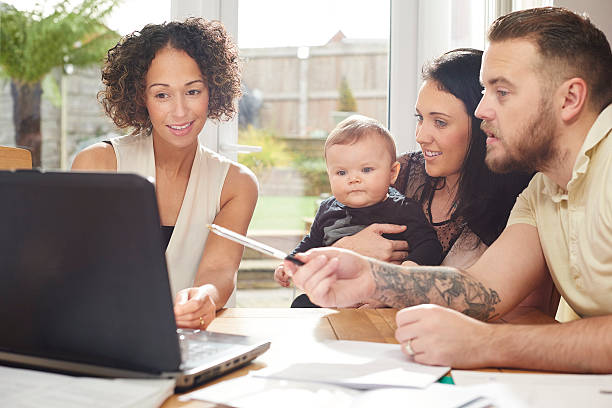 financial advisor with young family a young husband and wife with their baby sit and chat to a woman in their dining room . They are all referring to a laptop in front of them and various paperwork is dotted about the table. unknown gender stock pictures, royalty-free photos & images