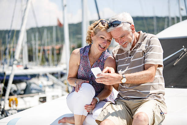 Senior couple having fun with smart phone on yacht The image shows a senior couple sailing and having fun on a yacht. They are sitting on a yacht, smiling and taking a selfie with a smartphone. In the background we can see the harbor and some boats. yacht selfie stock pictures, royalty-free photos & images