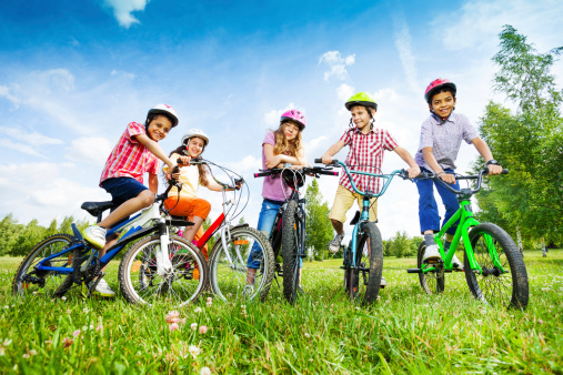 Children in colorful helmets hold bike handle-bars and are ready to ride their bikes in green field