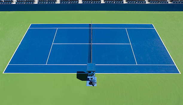 High angle view of a clean new professional tennis court.  Court is Blue surrounded by green.  Outdoor tennis court is a hard court with empty chair umpire seat. no tennis players on court photographed day time in sunlight.