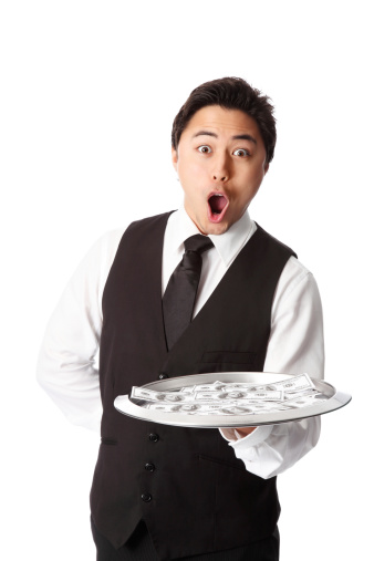 Attractive waiter in a vest and shirt, holding a tray with dollar bills. White background.