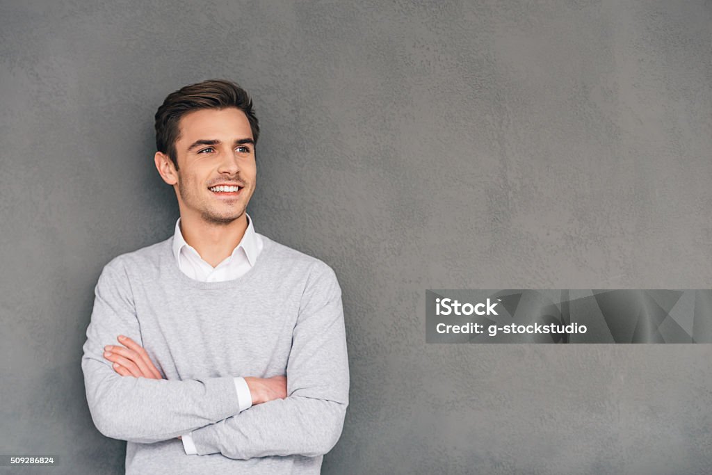 Looking in future with smile. Confident young man keeping arms crossed and looking away with smile while standing against grey background Men Stock Photo