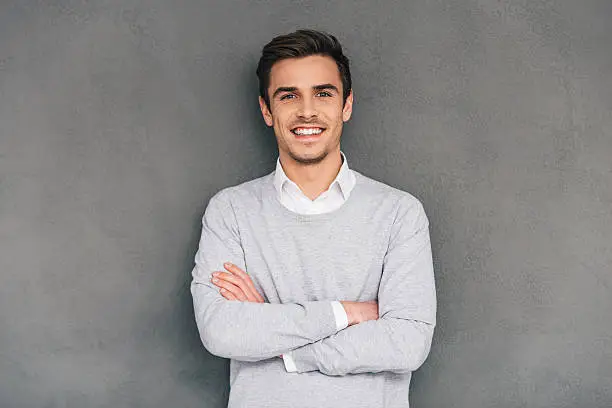 Confident young man keeping arms crossed and looking at camera with smile while standing against grey background