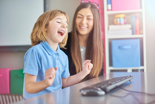 a primary school girl is sitting at her computer desk in school and doing a little desk dance of excitement at finishing her work on the computer . Her teacher looks on and laughs in the background .