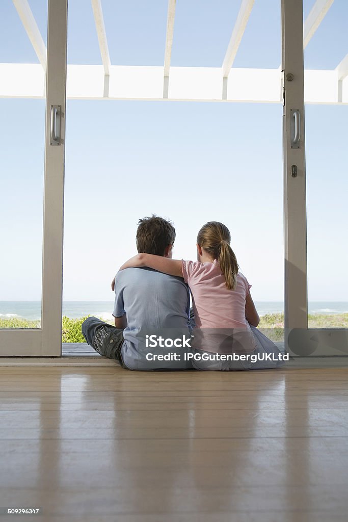 Siblings Sitting In Doorway Looking At View Rear view of young girl and boy sitting in doorway looking at view Arm Around Stock Photo