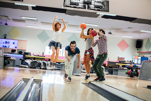 Group of happy friends having fun, bowling together. Front view of them bowling. Selective focus to young man in front just throw the ball. Friends in background jumping and cheering.