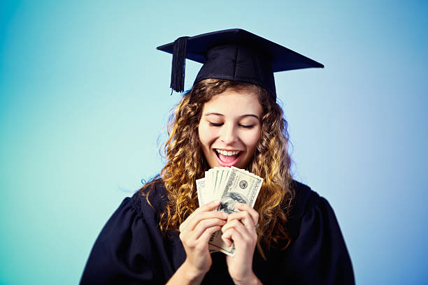 istock Pretty graduate looks down at bundle of banknotes, smiling 509346077