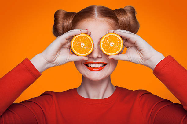 Young beautiful fashion model with orange. studio shot. Young beautiful funny fashion model with orange slice on orange background. with orange makeup and hairstyle and freckles. holding orange between eyes with toothy smile. artists model photos stock pictures, royalty-free photos & images