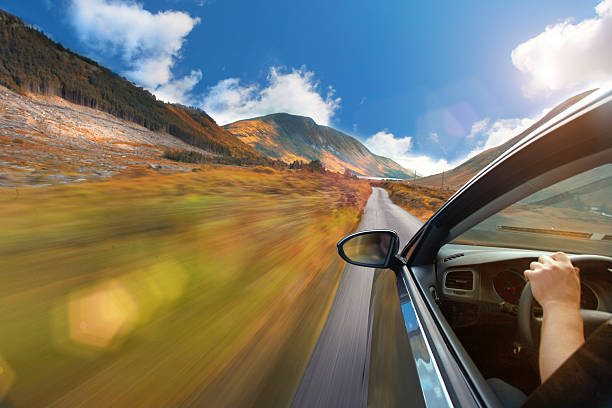 Car exterior, with window and rear-view mirror, while driving on a straight country road with a mountain landscape, surrounded by green fields. Driver's arm and hand on steering wheel are visible through the window, while conducting the car on the single lane narrow road. Blue sky with some clouds, illuminated by a bright sunshine creating a lens flare. Motion blur effect with sense of speed and freedom. Personal perspective, like watching out of the window. Copy space on left side of image.