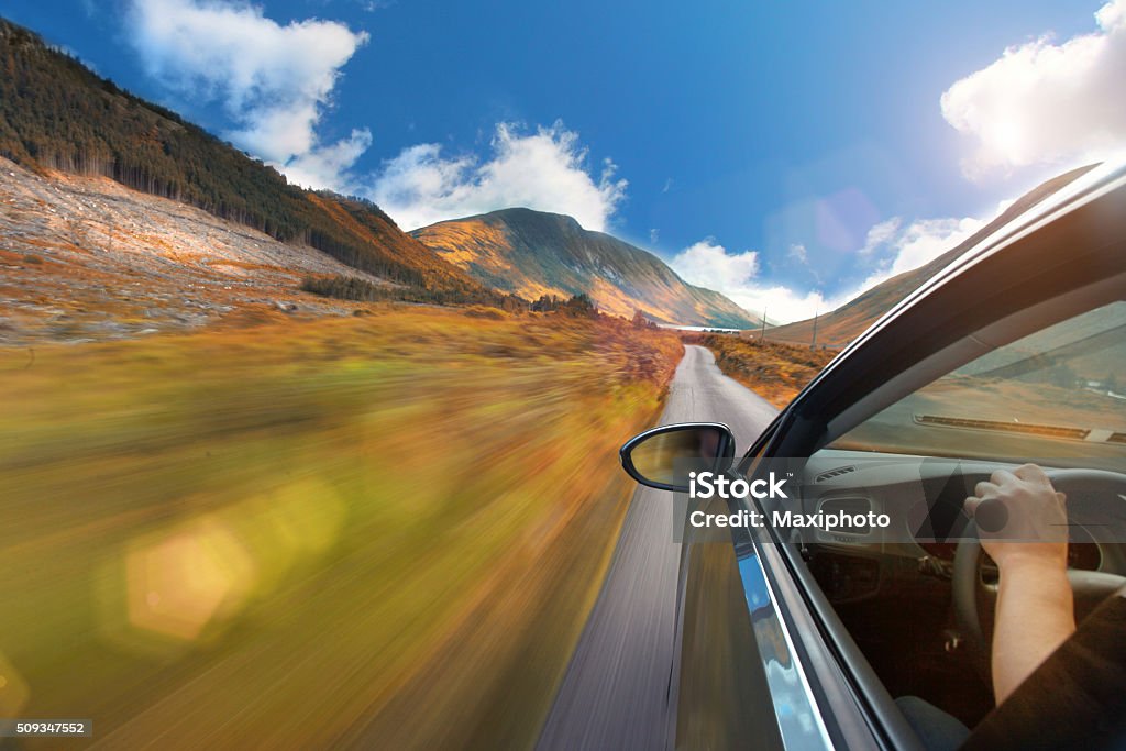 Driving a car on a mountain road with idyllic landscape Car exterior, with window and rear-view mirror, while driving on a straight country road with a mountain landscape, surrounded by green fields. Driver's arm and hand on steering wheel are visible through the window, while conducting the car on the single lane narrow road. Blue sky with some clouds, illuminated by a bright sunshine creating a lens flare. Motion blur effect with sense of speed and freedom. Personal perspective, like watching out of the window. Copy space on left side of image. Landscape - Scenery Stock Photo