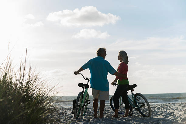 matura coppia con biciclette in spiaggia - mature adult bicycle senior adult heterosexual couple foto e immagini stock