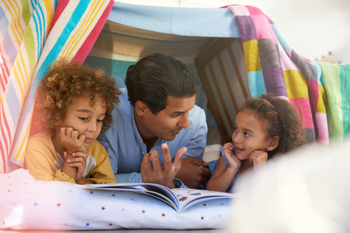 Shot of a young father lying in a tent fort with his children and reading them a book