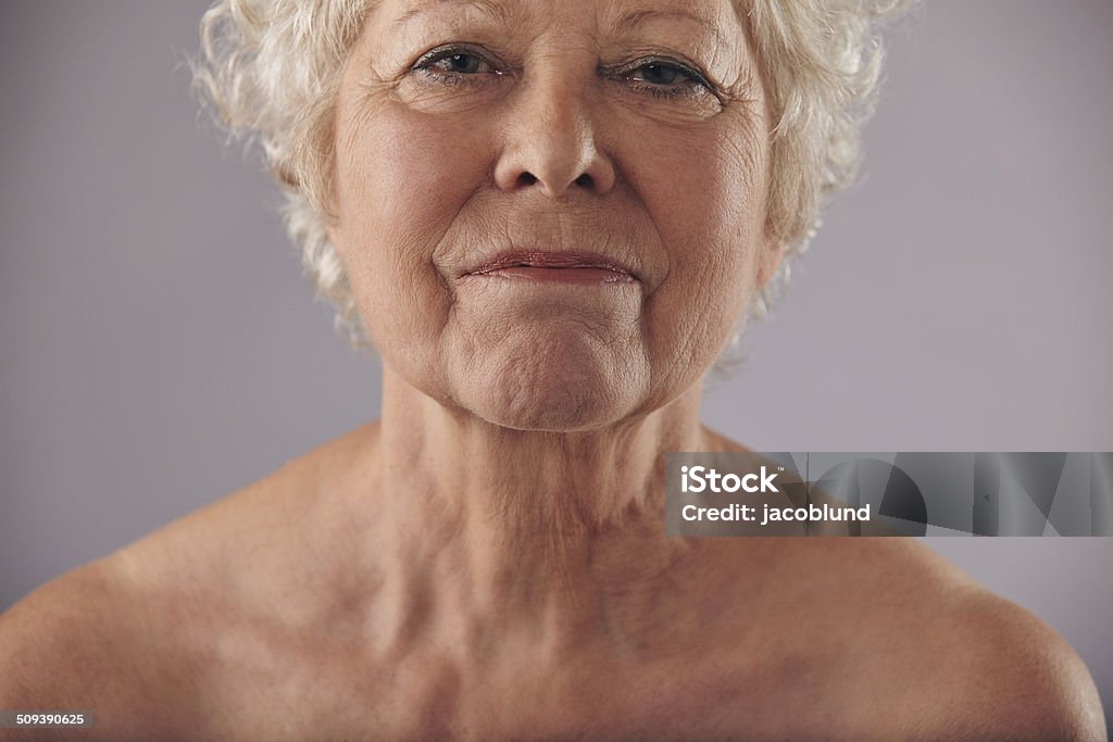 Mature woman face with wrinkled skin Close-up image of mature woman face with wrinkled skin condition against grey background. Senior woman naked chest and shoulders. Wrinkled Stock Photo