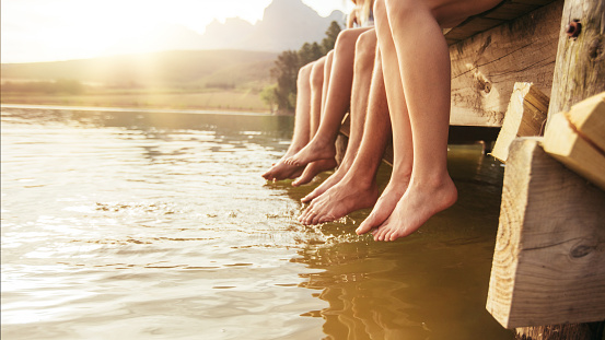 Four young friends sitting on jetting with their legs hanging down to the water on a summer day. Focus on legs of young people.