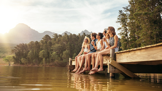 Portrait of relaxed young people sitting on the jetty with beers. Friends hanging out at the lake on a summer day.