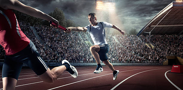 Running Pass on . Stadium A male athletes sprinting and passing race baton on the track on the . stadium. The bleachers are full of spectators. The sky is dark, cloudy and dramatic. Sportsmen are wearing an unbranded sport uniform. relay photos stock pictures, royalty-free photos & images