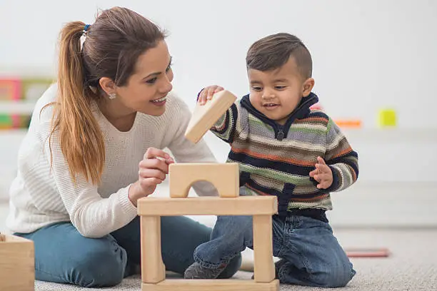 A mother and her toddler son are playing together in their home on mother's day. The little boy is building a block tower.