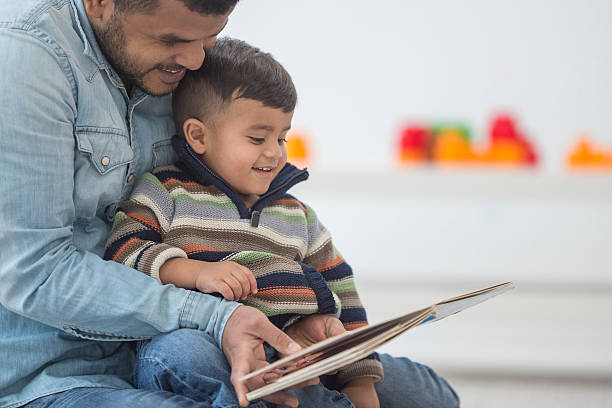 Father Reading His Son a Book A little boy is sitting in his dad's lap while his father reads him a book. They are both enjoying spending quality time together on father's day. toddler photos stock pictures, royalty-free photos & images