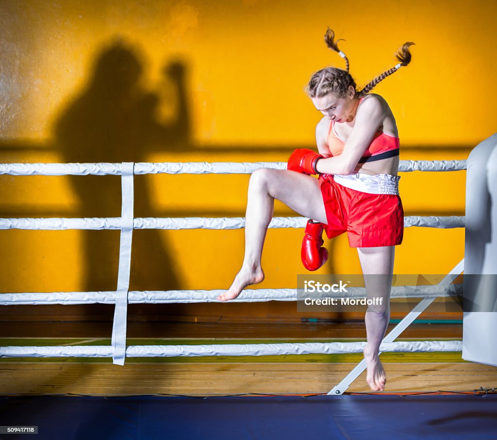 Doing Kickboxing Athletic girl teenager dressed in sports uniform during a fight kickboxing. The girl in boxing stance sideways to the camera. She is jumping for boxing kick to the shadow of man. Shooting in the gym Boxing - Sport Stock Photo