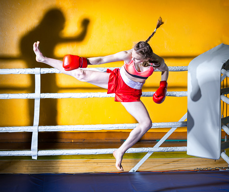 Athletic girl teenager dressed in sports uniform during a fight kickboxing. The girl in boxing stance sideways to the camera. She is jumping for boxing kick to the shadow of man. Shooting in the gym