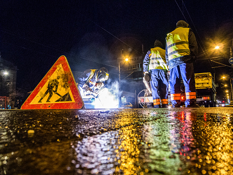 Low angle view of street workers and a welder while repairing the rail tracks in the city at night. Road sign is in front and truck with equipment is in the background.