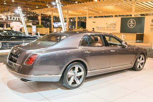 Brussels, Belgium - Januari 12, 2016: Gray Bentley Mulsanne Speed luxury sedan luxury sedan rear view. The Speed model offers even greater performance and more premium features compared to a standard Bentley Mulsanne. The Mulsanne Speed is equipped 6.75-litre twin-turbocharged V8 petrol engine that produces 530 bhp. The car is on display during the 2016 Brussels Motor Show. The car is displayed on a motor show stand, with lights reflecting off of the body. There are people looking around and other cars on display in the background.