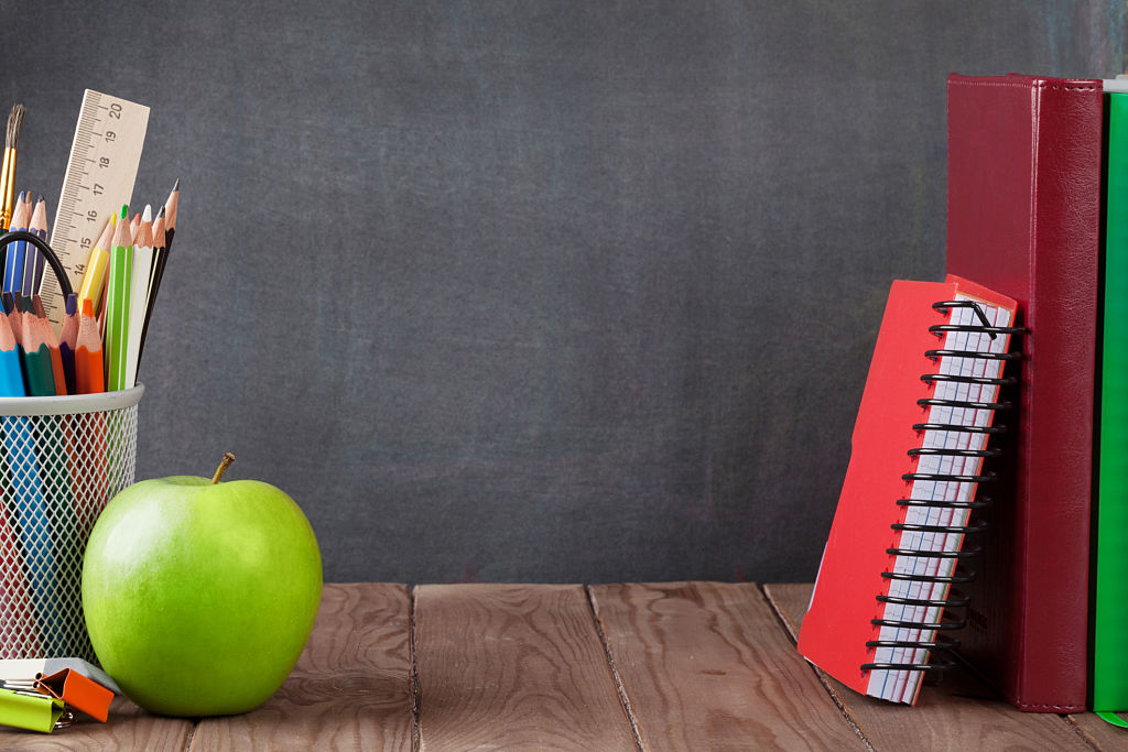 Schul- und Bürobedarf und Apfel auf dem Klassenzimmertisch vor der Tafel. Ansicht mit Kopierbereich
