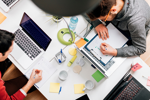 Young programmers working in startup company. Working from the house like business incubator.  Top view of men using his laptops for coding and writing. Coworkers sitting by the table and working.