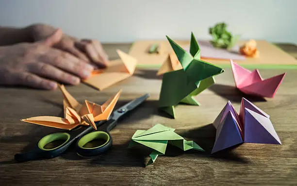 A few origami figurines on the wooden table, in the background hands folding colored paper.